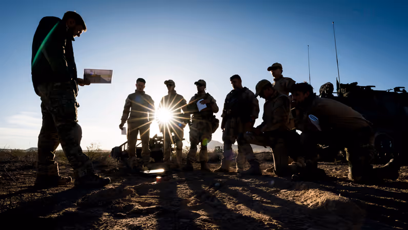 Een groep militairen kijkt naar een militair voor hen die iets uit zijn boekje leest. Van hen zijn, door de zon achter hen, enkel silhouetten te zien. 