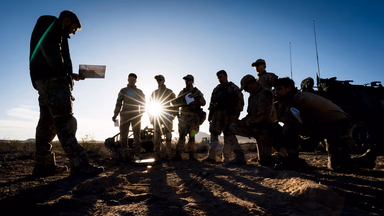 Een groep militairen kijkt naar een militair voor hen die iets uit zijn boekje leest. Van hen zijn, door de zon achter hen, enkel silhouetten te zien. 