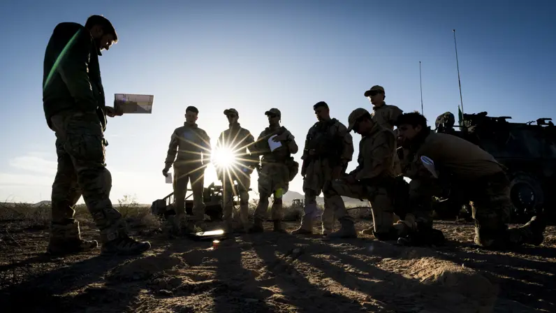 Een groep militairen kijkt naar een militair voor hen die iets uit zijn boekje leest. Van hen zijn, door de zon achter hen, enkel silhouetten te zien. 