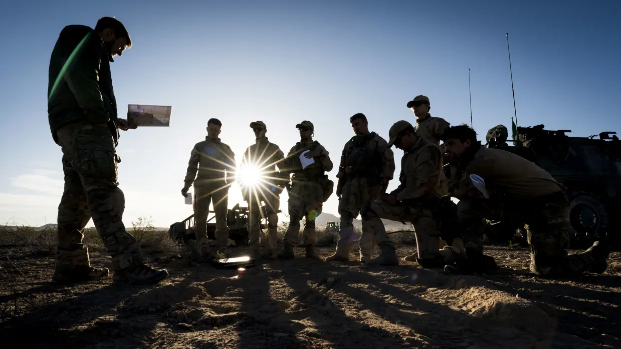 Een groep militairen kijkt naar een militair voor hen die iets uit zijn boekje leest. Van hen zijn, door de zon achter hen, enkel silhouetten te zien. 
