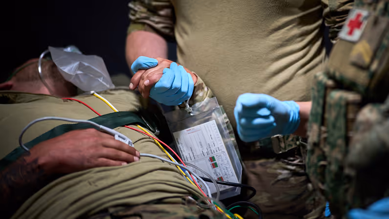 Een gewonde militair ligt op een bed. Een andere militair houdt zijn hand vast. Hij draagt een blauwe rubberen handschoen. 