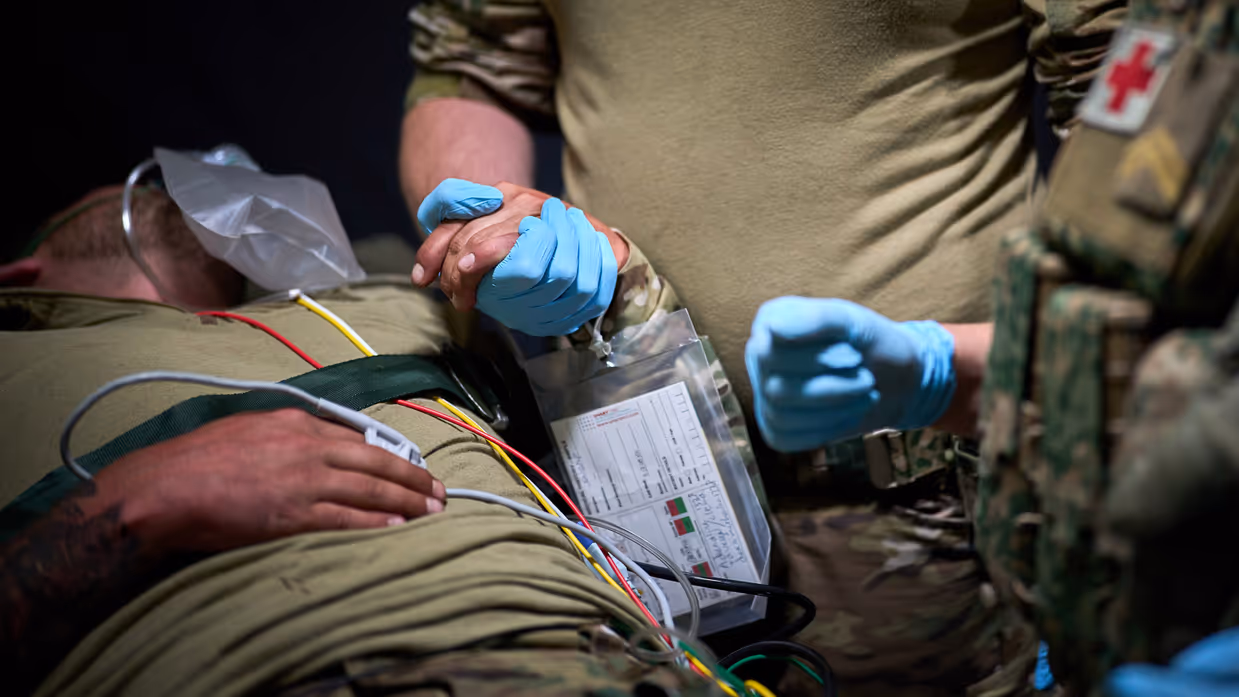 Een gewonde militair ligt op een bed. Een andere militair houdt zijn hand vast. Hij draagt een blauwe rubberen handschoen. 