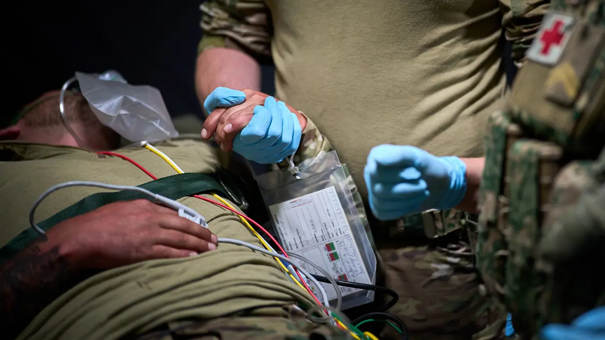 Een gewonde militair ligt op een bed. Een andere militair houdt zijn hand vast. Hij draagt een blauwe rubberen handschoen. 