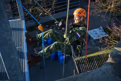 Koningin Maxima hangt aan touwen tijdens een oefening en heeft een oranje helm op.
