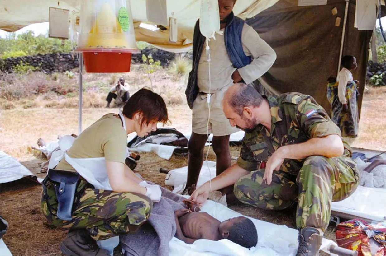 2 militairen van een medisch team verzorgen een vluchteling die met een infuus op een brancard ligt in een vluchtelingenkamp.
