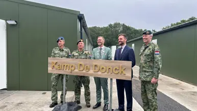Drie militairen, de staatssecretaris en een burgemeester achter een houten bord met naam ‘Kamp De Donck’. 