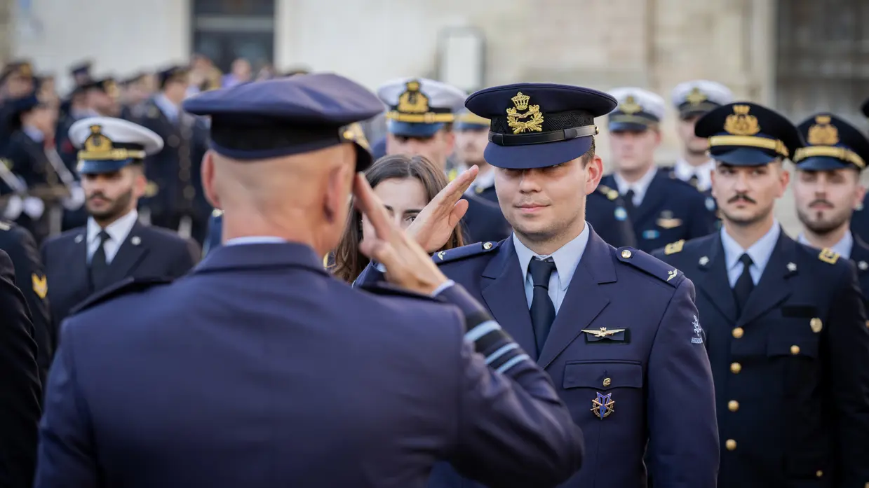 Steur en een leerling-vlieger met wing brengen elkaar de militaire groet. Achter de leerling staat een groep militairen opgesteld uit verschillende landen. 