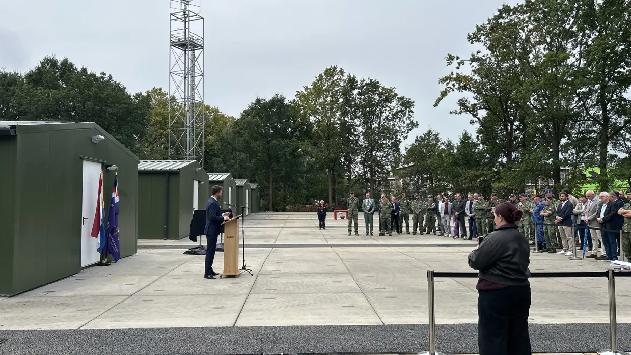De staatssecretaris spreekt vanachter een spreekgestoelte op een terrein met achter hem vijf legergroene prefab-gebouwen. Voor hem staat in een brede boog publiek, bestaande uit militairen en burgers.