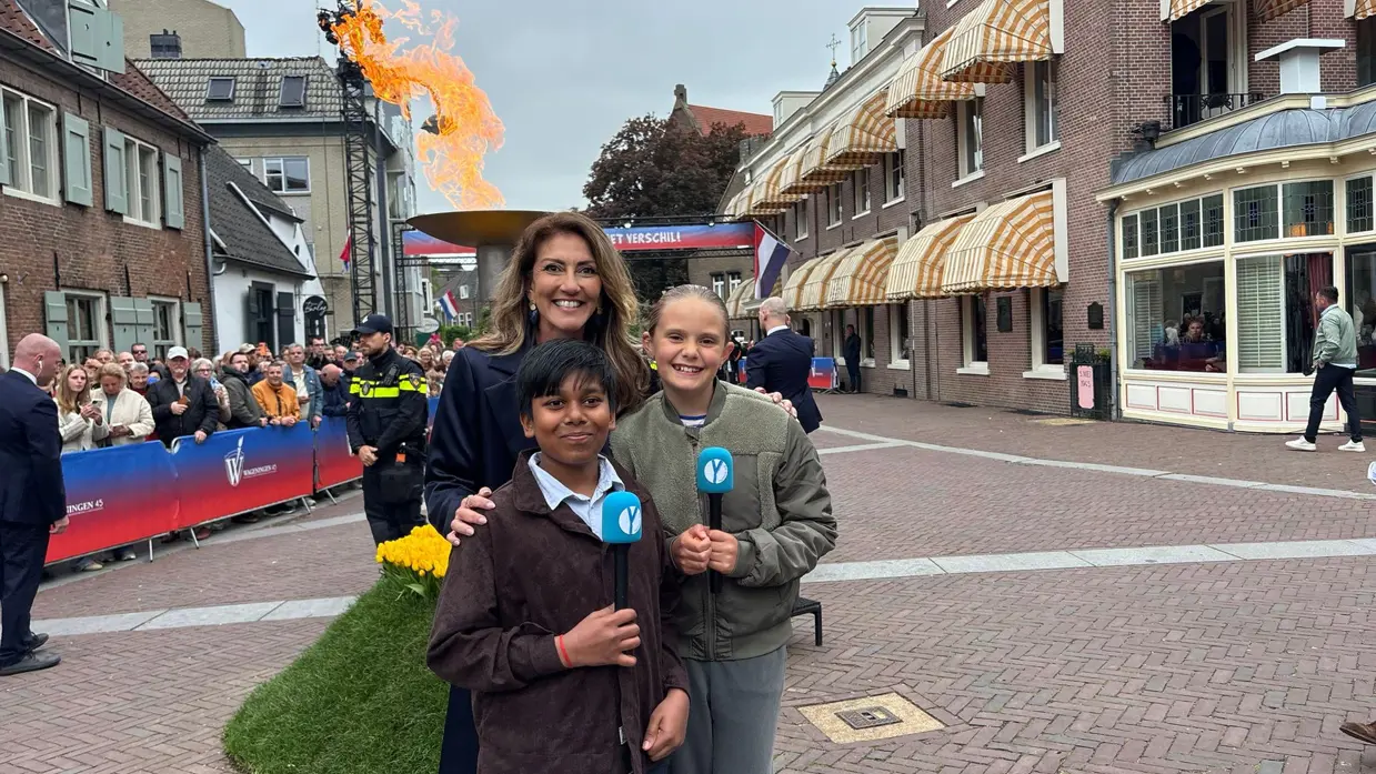 Minister Dilan Yeşilgöz met twee kindercorrespondenten met microfoons. Op de achtergrond het bevrijdingsvuur.