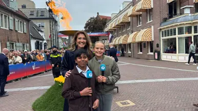 Minister Dilan Yeşilgöz met twee kindercorrespondenten met microfoons. Op de achtergrond het bevrijdingsvuur.