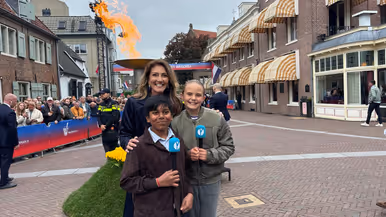 Minister Dilan Yeşilgöz met twee kindercorrespondenten met microfoons. Op de achtergrond het bevrijdingsvuur.