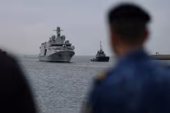 Het grijze schip De Vlissingen komt aangevaren. Rechts vaag een marinier in beeld.