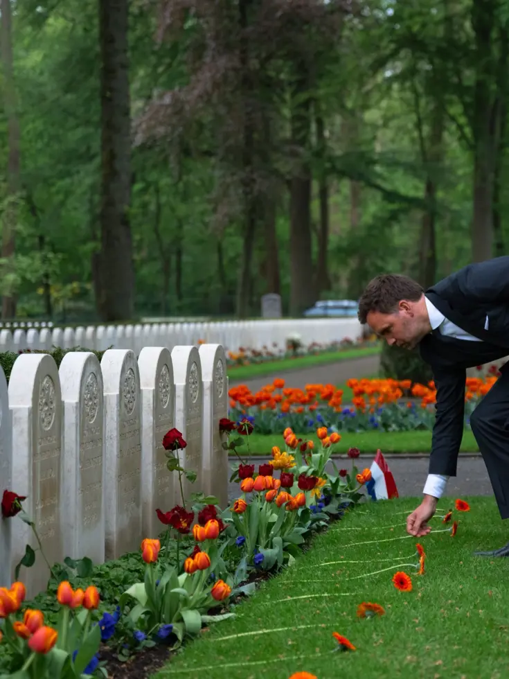 Staatssecretaris Derk Boswijk legt een oranje bloem op een oorlogsgraf met witte grafsteen op het Ereveld Grebbeberg. Op de andere graven liggen dezelfde oranje bloemen.
