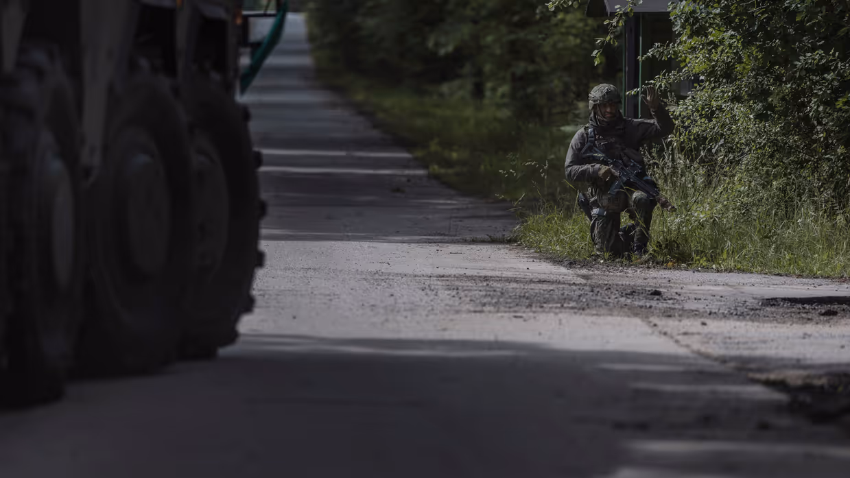 Militair in groen tenue aan de kant van de weg met zijn hand omhoog.