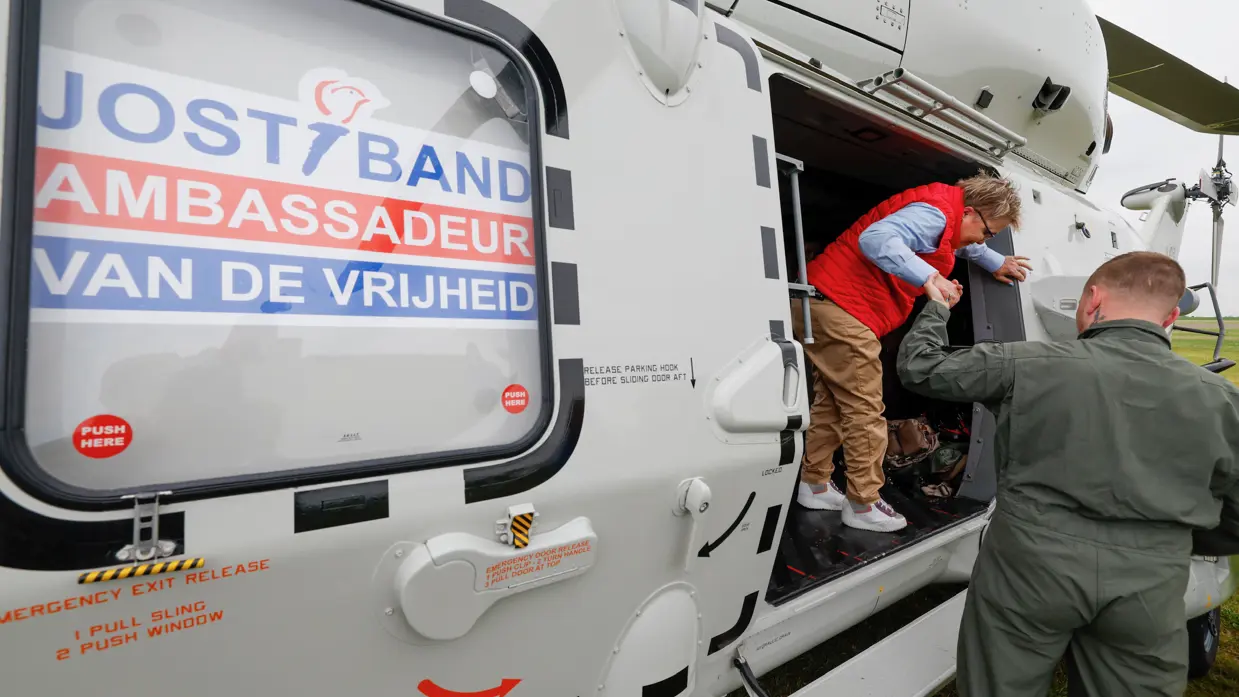 Een militair helpt een lid van de Jostiband uit de NH90 stappen. Op de helikopter staat de tekst ‘Jostiband, Ambassadeur van de Vrijheid’.