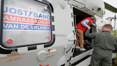 Een militair helpt een lid van de Jostiband uit de NH90 stappen. Op de helikopter staat de tekst ‘Jostiband, Ambassadeur van de Vrijheid’.