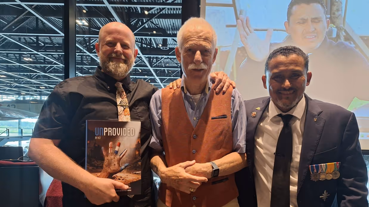Drie mannen staan samen, een houdt het boek "UNPROVIDED" vast, achtergrond met vliegtuigen en een groot scherm. 
