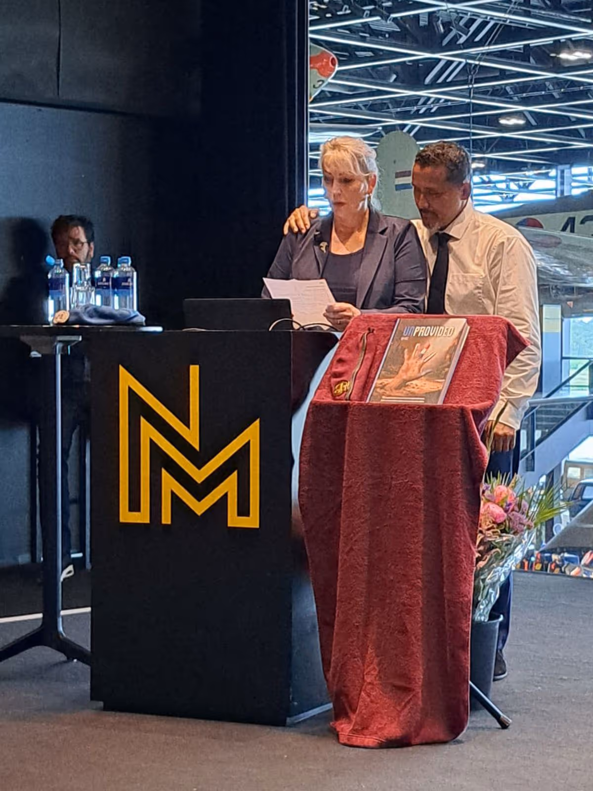Een vrouw en man staan achter een podium met een rode doek, microfoon en bloemen. Achtergrond: vliegtuig en NMM-logo.