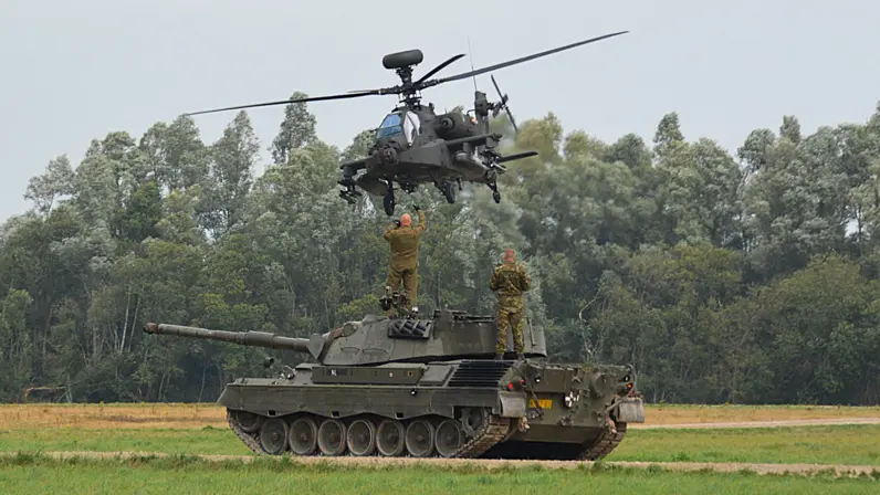 Een gevechtshelikopter zweeft boven een tank in een groen veld, met twee soldaten op de tank en bomen op de achtergrond.