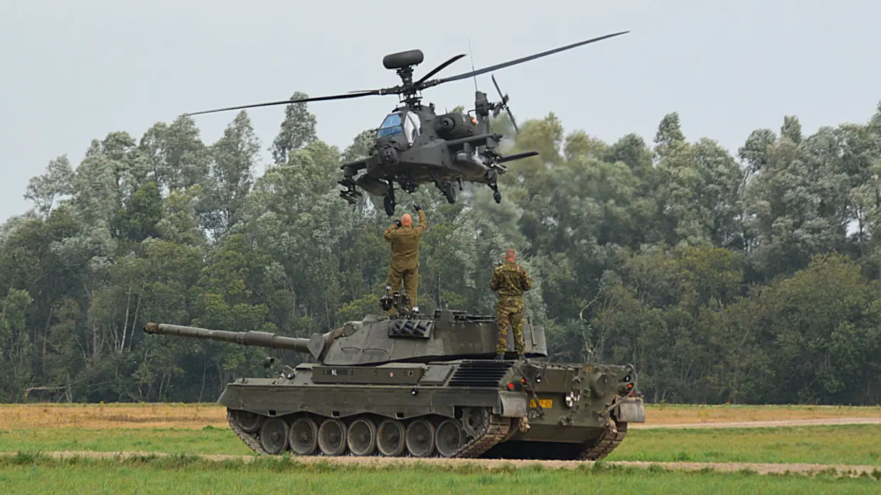 Een gevechtshelikopter zweeft boven een tank in een groen veld, met twee soldaten op de tank en bomen op de achtergrond.