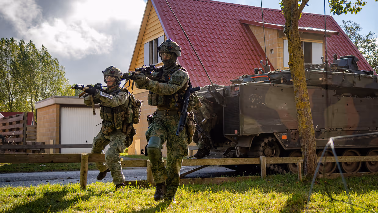 Twee militairen lopen met getrokken geweren rond, met een voertuig en huis in de achtergrond.