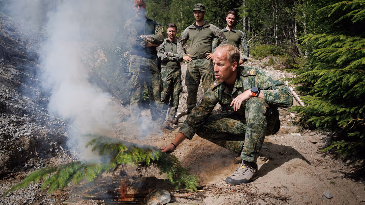 Een groep militairen in een Noorweegs bos, op de voorgrond is een militair een vuurtje aan het stoken.