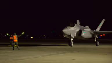 In het donker loodst grondpersoneel met een oranje hesje aan en met lichtgevende sticks in de handen een F-35 naar zijn parkeerplaats.