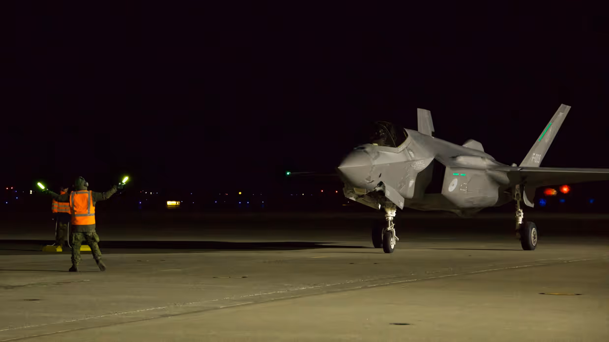 In het donker loodst grondpersoneel met een oranje hesje aan en met lichtgevende sticks in de handen een F-35 naar zijn parkeerplaats.