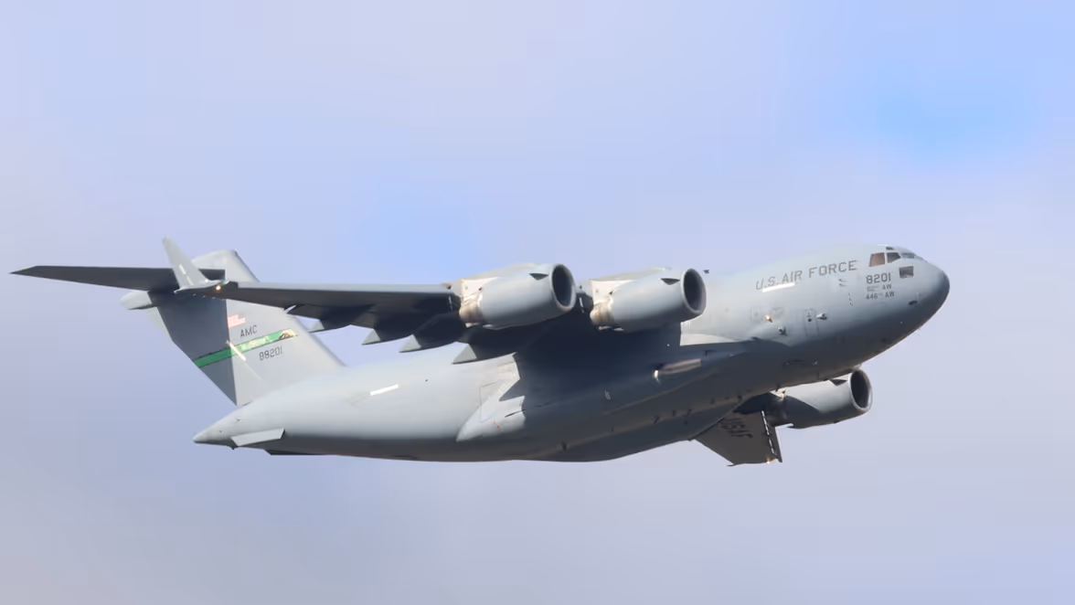 Een groot grijs militair vliegtuig vliegt onder een blauwe lucht. 