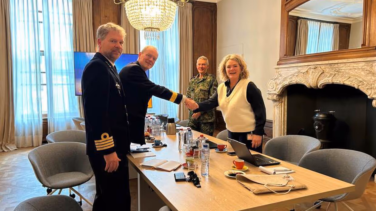 3 militairen in uniform en 1 vrouw staan rond een tafel. De vrouw in burgerkleding en een militair schudden de hand.