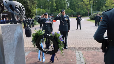 Twee militairen in uniform salueren bij een krans met bloemen en linten, omringd door andere militairen in een groene, open omgeving. 