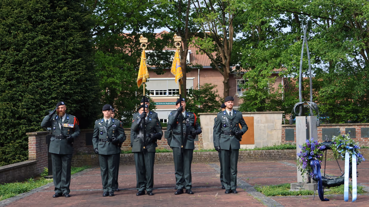 Vijf militairen in groene uniformen salueren in een rij, met vlaggen en een krans met bloemen op een stenen plein.