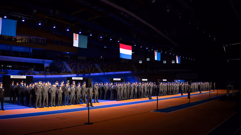 Militairen in dagelijks tenue staan naast elkaar opgesteld in een grote hal tijdens medaille-uitreiking.
