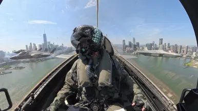 Een F-35-vlieger frontaal in beeld binnenin zijn cockpit. Links van het toestel vliegen nog twee F-35’s en rechts ook een. Ze vliegen over een rivier met links en rechts daarvan de skyline van New York.