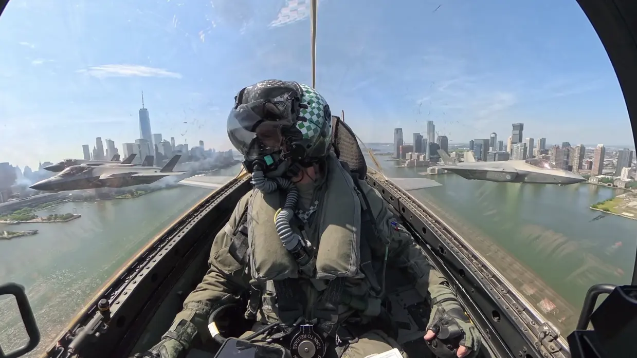 Een F-35-vlieger frontaal in beeld binnenin zijn cockpit. Links van het toestel vliegen nog twee F-35’s en rechts ook een. Ze vliegen over een rivier met links en rechts daarvan de skyline van New York.