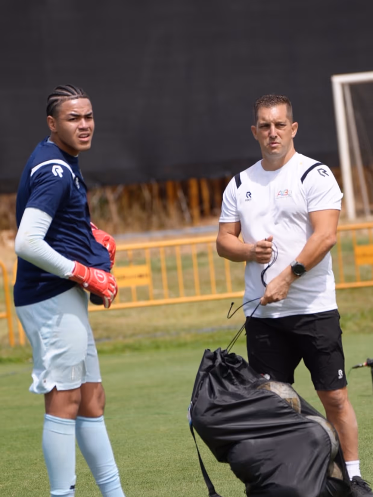 Een keeper met keepershandschoenen aan en een trainer met de ballentas. 