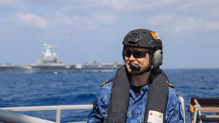 Alttekst: LTZ 2 (LD) Thom Cardol op de voorgrond met helm, zwemvest en zonnebril. Op de achtergrond de carrier Charles de Gaulle.