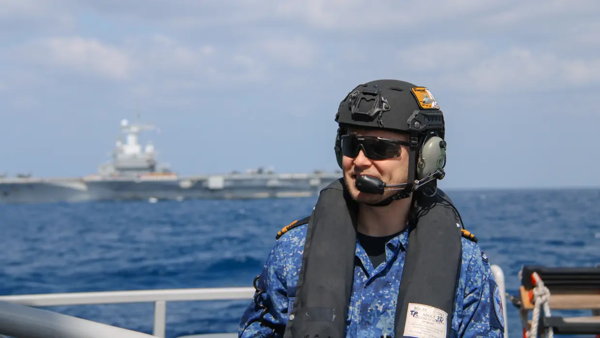 Alttekst: LTZ 2 (LD) Thom Cardol op de voorgrond met helm, zwemvest en zonnebril. Op de achtergrond de carrier Charles de Gaulle.