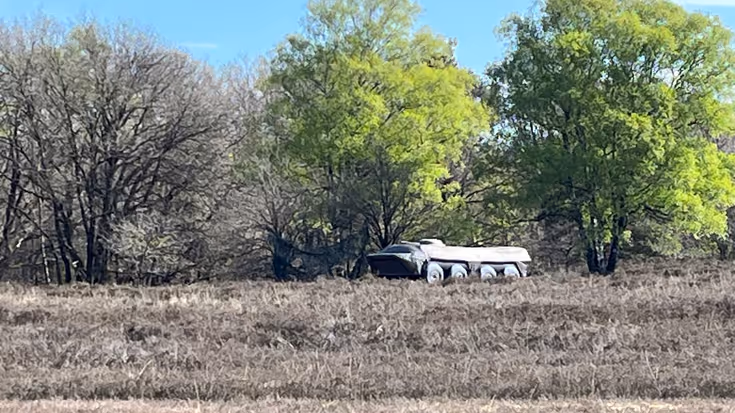 Met bosschages op de achtergrond staat een groen lang voertuig op een zandpad. Het is zonnig weer.