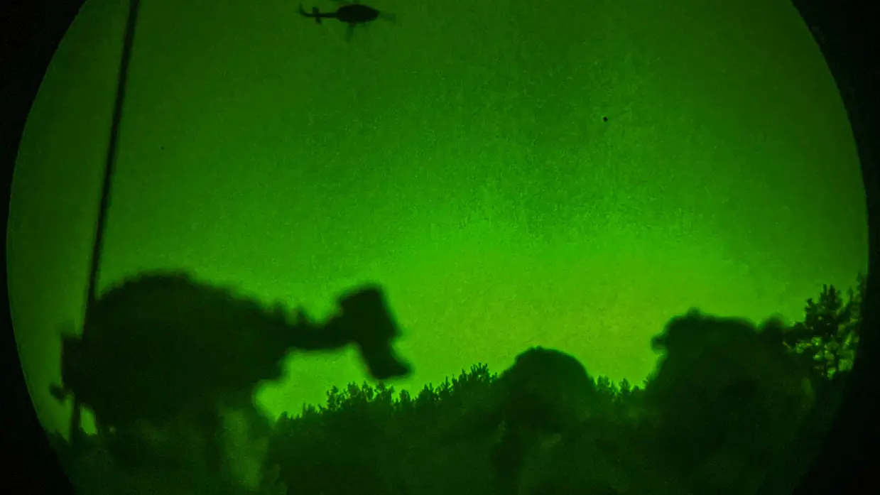 Een groep wachtende militairen zijn zichtbaar door een nachtlens, boven in de lucht vliegt een helikopter.