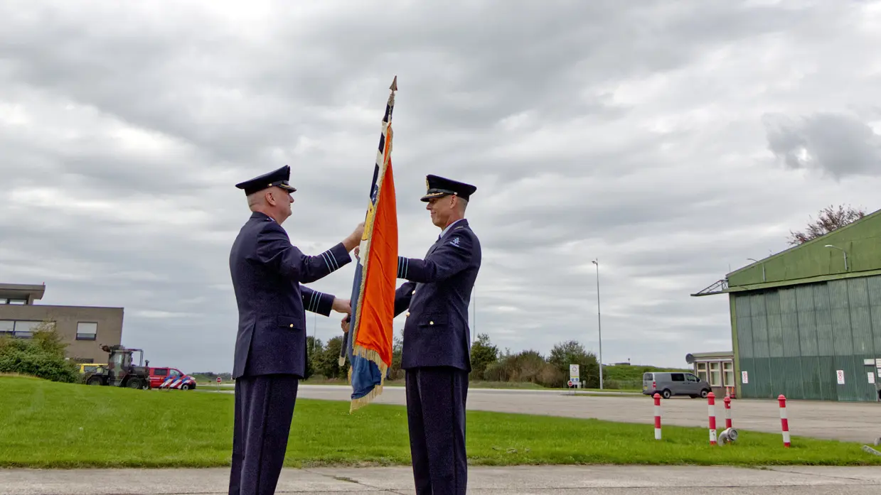 Twee militairen die elkaar een vlag overhandigen. Erboven vliegt een MQ-9 Reaper tegen een grijs bewolkte lucht.