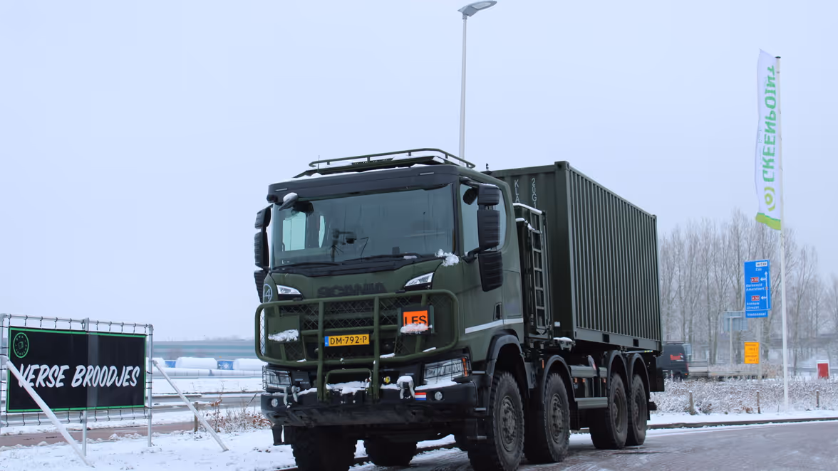 Een groot, groen militair voertuig met een gesloten container staat stil op een besneeuwde weg langs een bord met opschrift ‘verse broodjes’ en een verkeersbord richting Vaassen.
