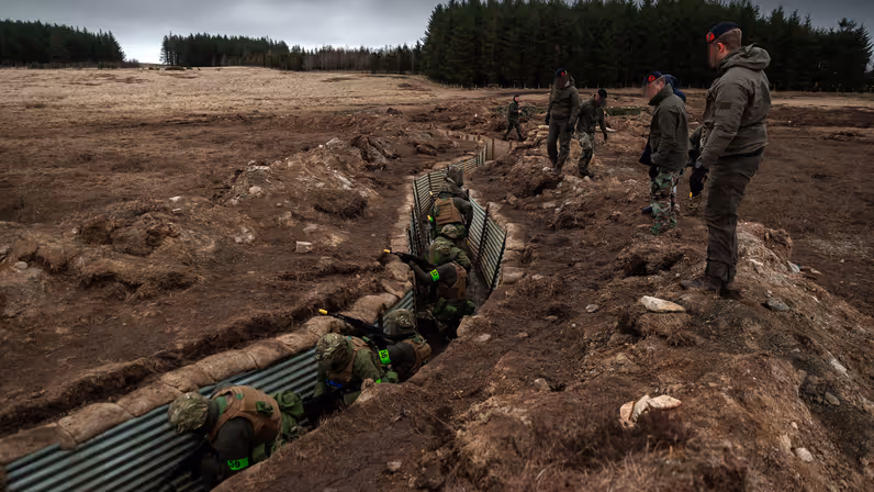 Alt-tekst: Militairen verplaatsen zich achter elkaar door een loopgraaf. Van bovenaf worden zij geobserveerd door instructeurs. 
