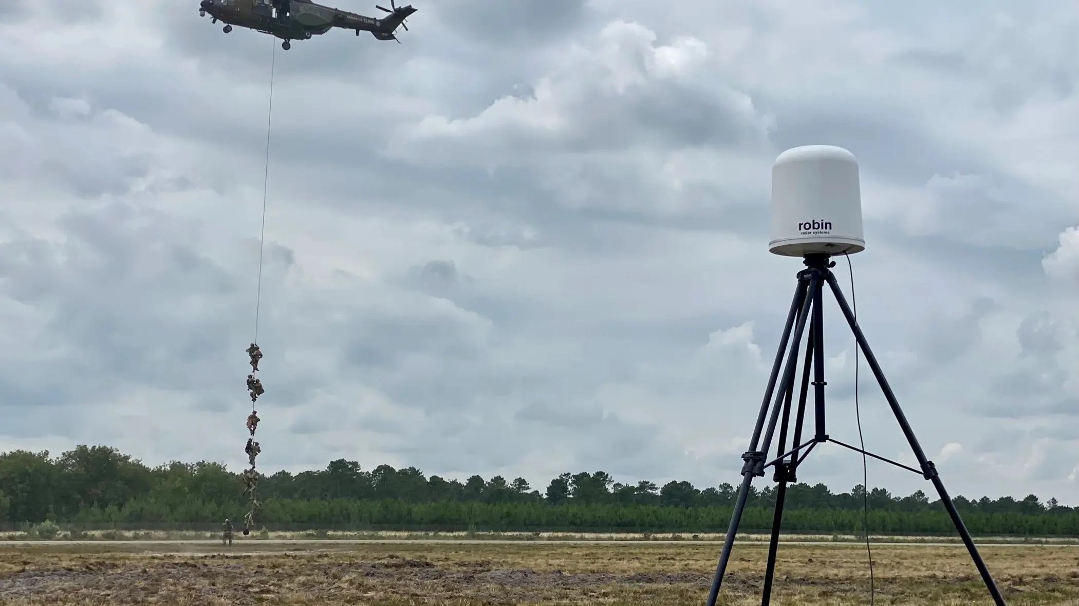Op de voorgrond een radar, op de achtergrond militairen die met een touw afdalen uit een helikopter. 