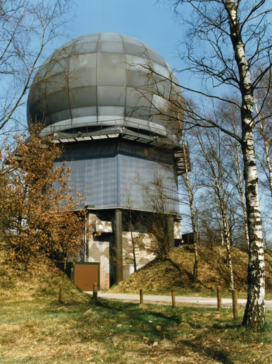 Een radarbol op een laag gebouw met links begroeiing en rechts een boom. 