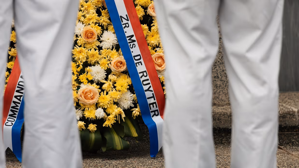 Militairen staan in tropentenue opgesteld op een militair ereveld in Soerabaja, op Java, en brengen de militaire groet. Op de andere foto staat de krans met opdruk Zr.Ms. De Ruyter die zij net hebben gelegd.  &nbsp;