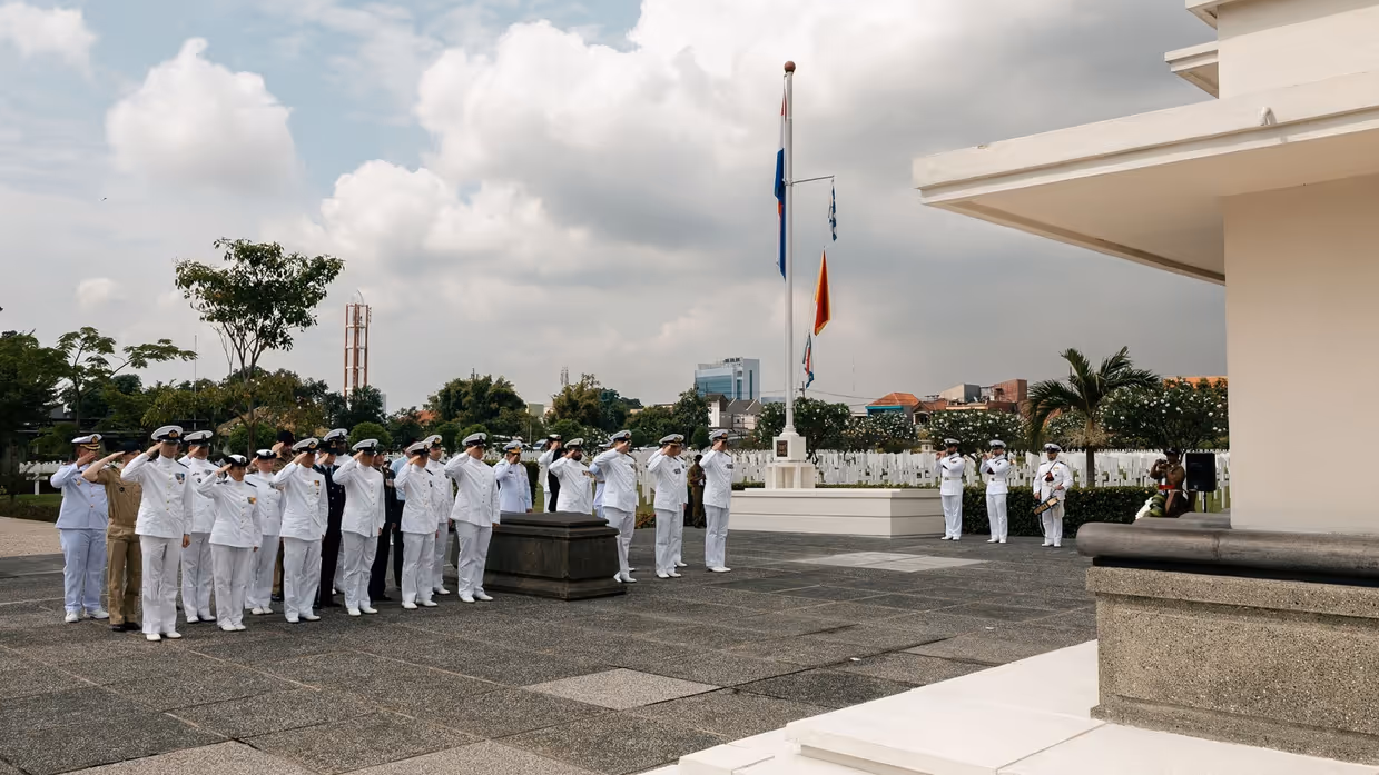 Militairen staan in tropentenue opgesteld op een militair ereveld in Soerabaja, op Java, en brengen de militaire groet. Op de andere foto staat de krans met opdruk Zr.Ms. De Ruyter die zij net hebben gelegd.