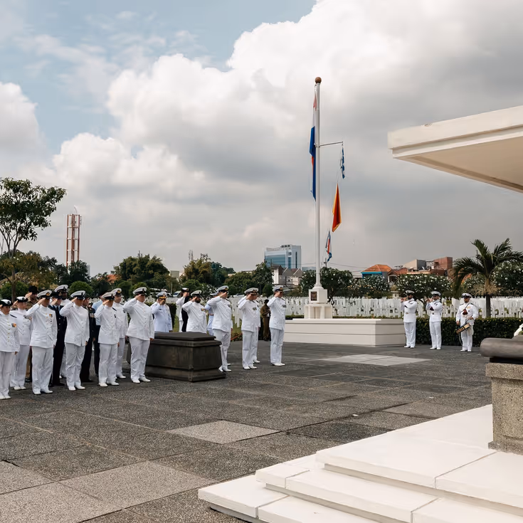 Militairen staan in tropentenue opgesteld op een militair ereveld in Soerabaja, op Java, en brengen de militaire groet. Op de andere foto staat de krans met opdruk Zr.Ms. De Ruyter die zij net hebben gelegd.