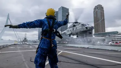 Een persoon in een blauw vlekkenpak met een gele helm op gebaart op een helikopterdek van een marineschip naar een vlak boven het dek vliegende NH90. Op de achtergrond de skyline van Rotterdam.