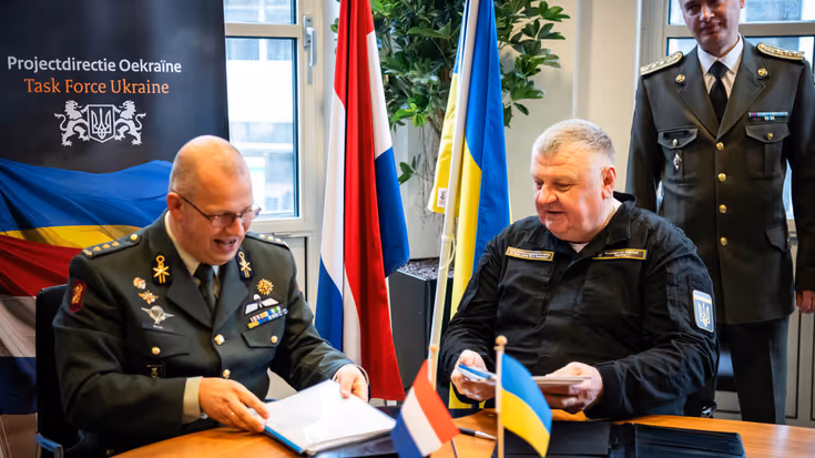 Kolonel Simon Wouda en chief engineer Vadym Nurmahomedov tekenen zittend aan een tafel de overdracht. Achter hen de Nederlandse en Oekraïense vlag.